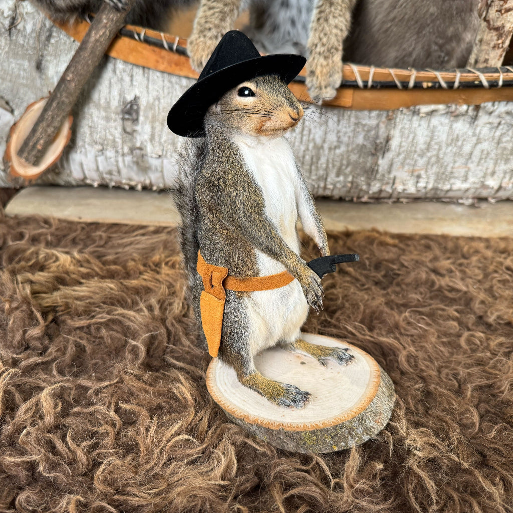 Squirrel figurine wearing a cowboy hat and bandana on a wooden platform with a rustic background.
